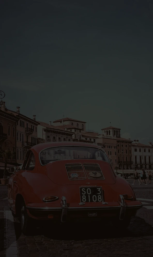 A photo of a red car with old buildings in the background, captured in Verona, Italy. The image has a CSS filter with 20% brightness, making it significantly less bright, almost black, compared to the original.